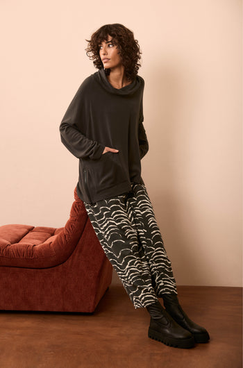Wearing Etched Wave Jersey Bubble Trousers with a dark cowl-neck sweater and black boots, a person leans against a rust-colored chaise lounge on a brown floor with a beige wall.
