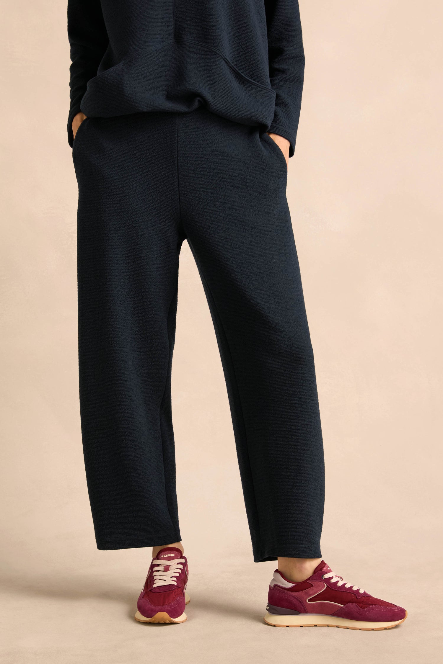 A person is wearing the Double Jersey Crinkle Trousers with a black sweatshirt, hands in pockets, maroon sneakers, and standing on a beige background. These trousers are made in England.