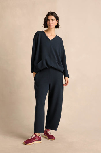 Woman standing against a beige background, wearing relaxed leg Double Jersey Crinkle Trousers in navy with a loose-fitting v-neck top and red sneakers.