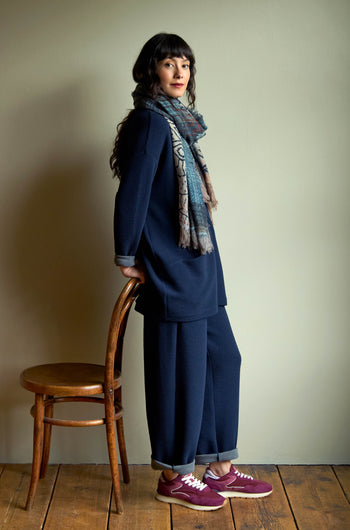 A woman in a dark blue top and matching Double Jersey Crinkle Trousers stands leaning against a wooden chair, accessorized with a patterned scarf and burgundy sneakers.