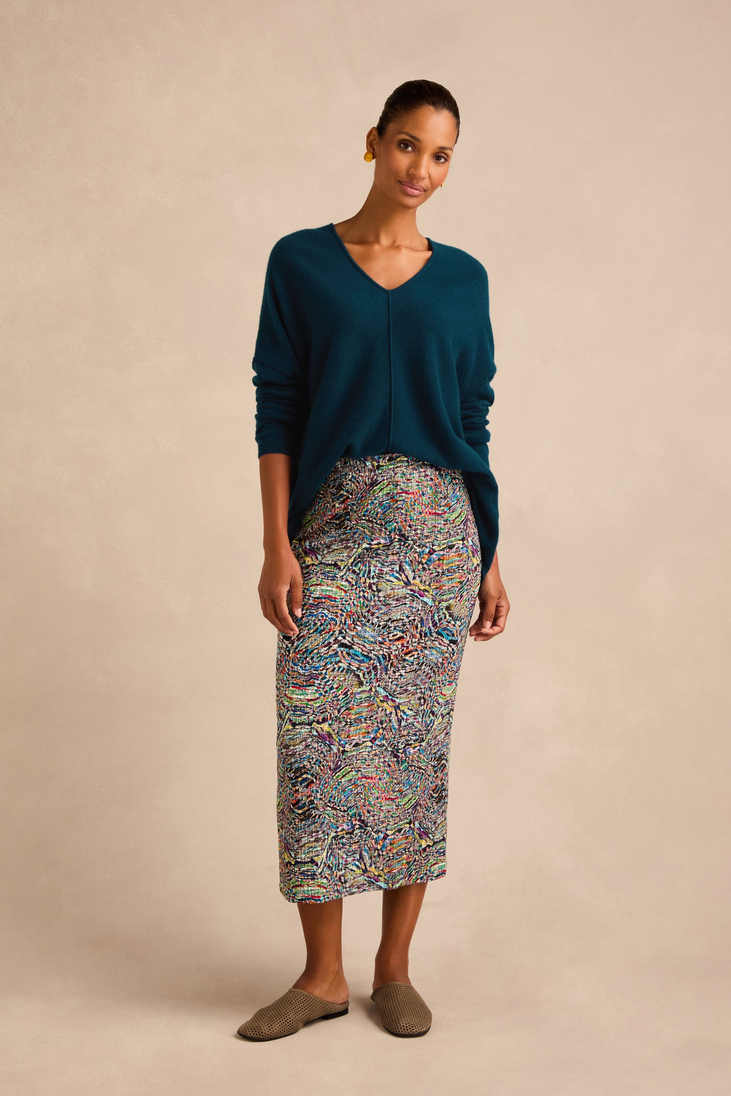 A woman stands against a beige background wearing a teal V-neck sweater, the Patchwork Fields Jersey Tube Skirt with colorful patterns, brown slip-on shoes, and gold earrings.