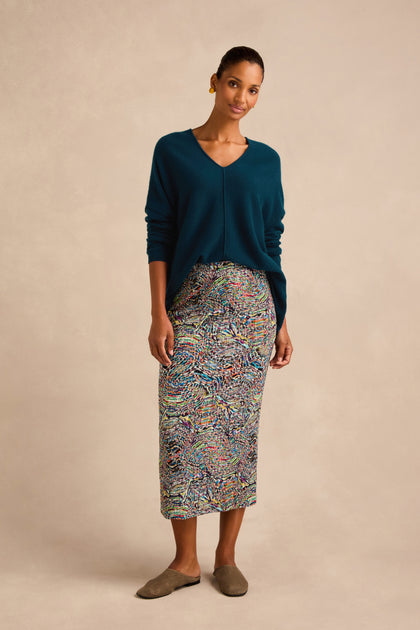 A woman stands against a beige background wearing a teal V-neck sweater, the Patchwork Fields Jersey Tube Skirt with colorful patterns, brown slip-on shoes, and gold earrings.
