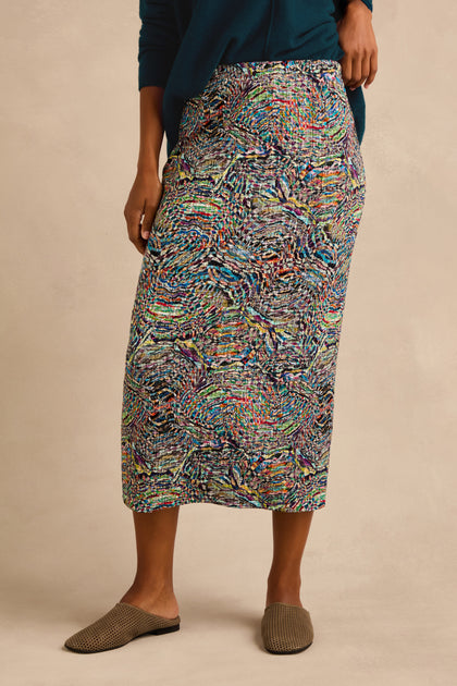 Wearing the Patchwork Fields Jersey Tube Skirt with a blue top and brown woven slip-on shoes, the person stands against a plain background.