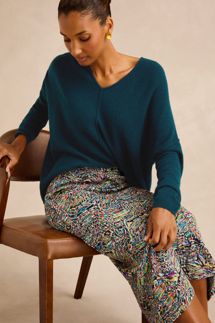 Woman seated on a brown chair wearing a teal V-neck sweater, the Patchwork Fields Jersey Tube Skirt with multicolored prints, and gold earrings, looking down with one hand resting on her knee.