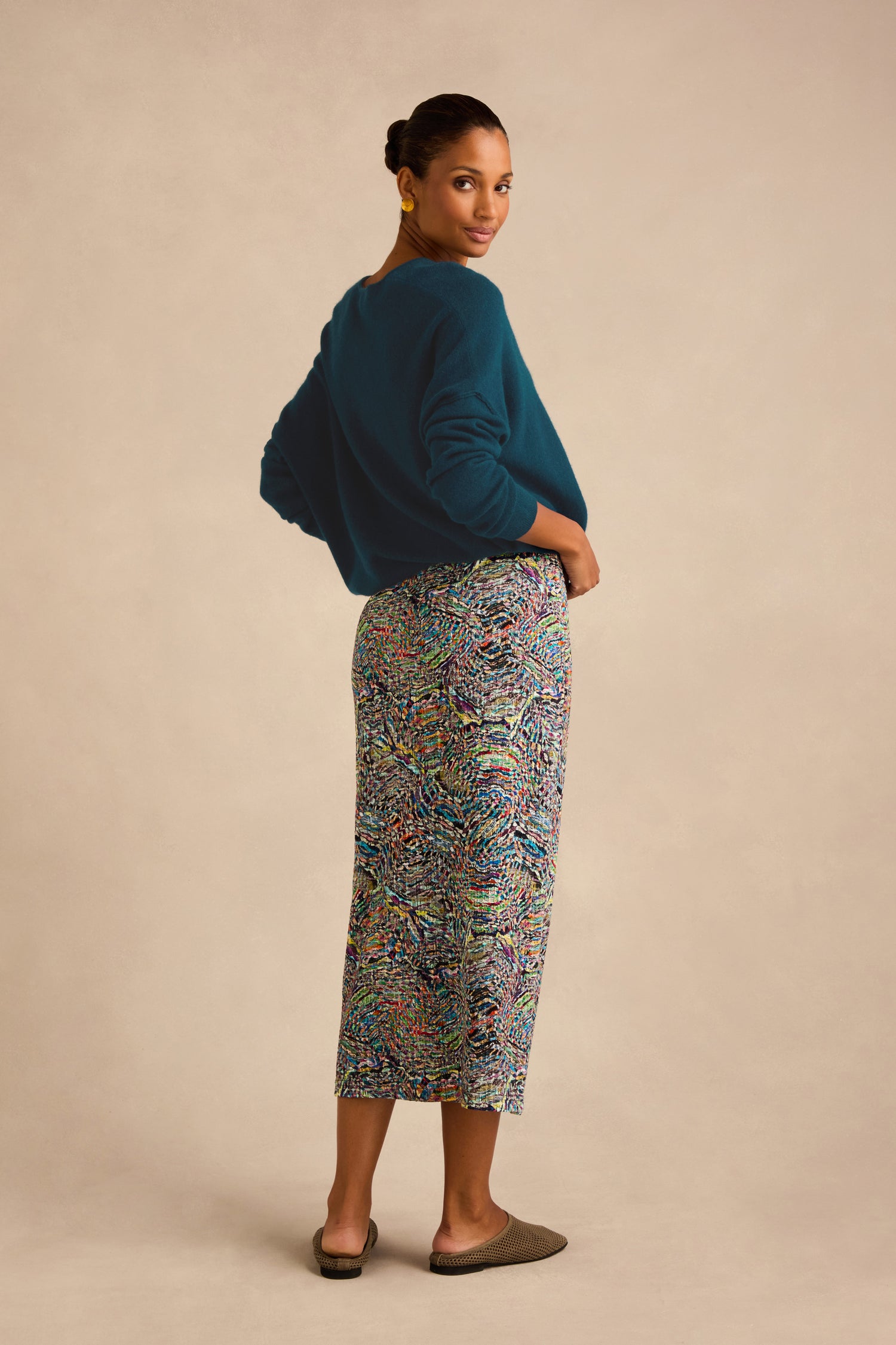 A woman stands sideways, looking over her shoulder, wearing a teal sweater, the Patchwork Fields Jersey Tube Skirt, and brown slip-on shoes against a neutral background.