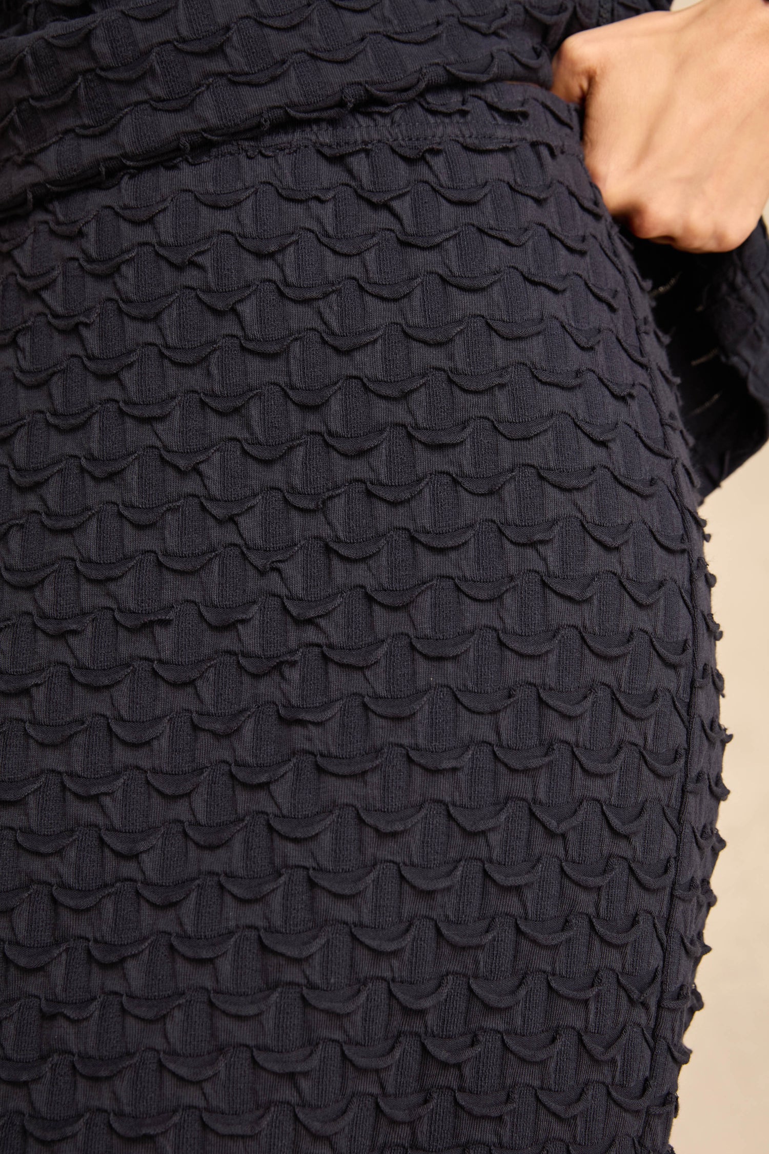 Close-up of a person wearing the Pocket Jersey Tube Skirt in a dark textured finish with a repeating scalloped pattern, hand resting near the waistband.