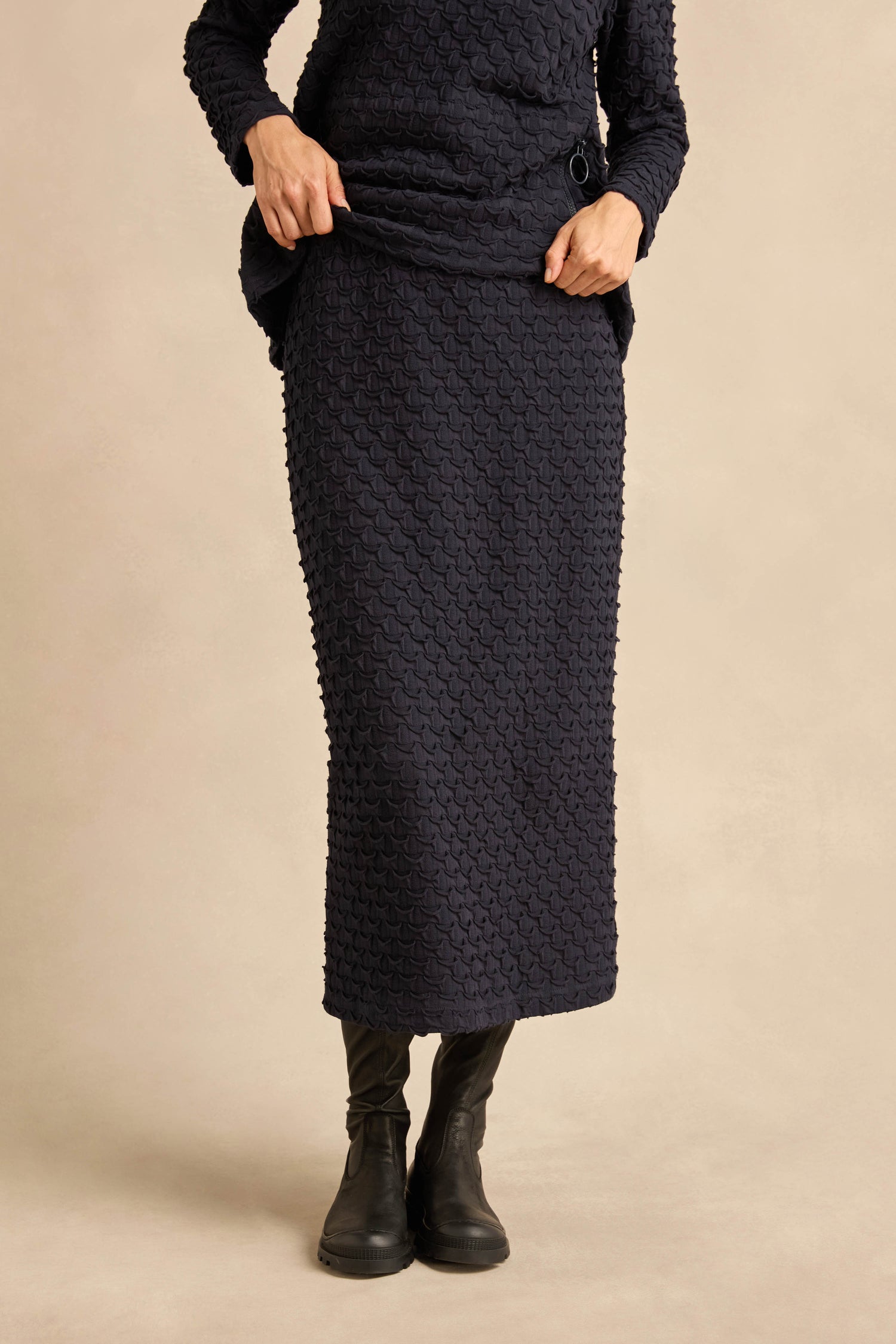 A person is wearing a textured navy long-sleeve top paired with the Pocket Jersey Tube Skirt in matching textured stretch fabric and black boots, standing against a plain beige background.