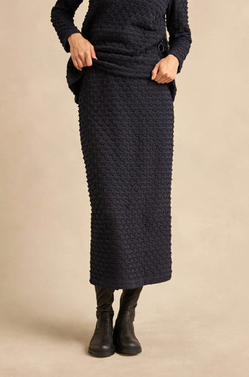 A person is wearing a textured navy long-sleeve top paired with the Pocket Jersey Tube Skirt in matching textured stretch fabric and black boots, standing against a plain beige background.