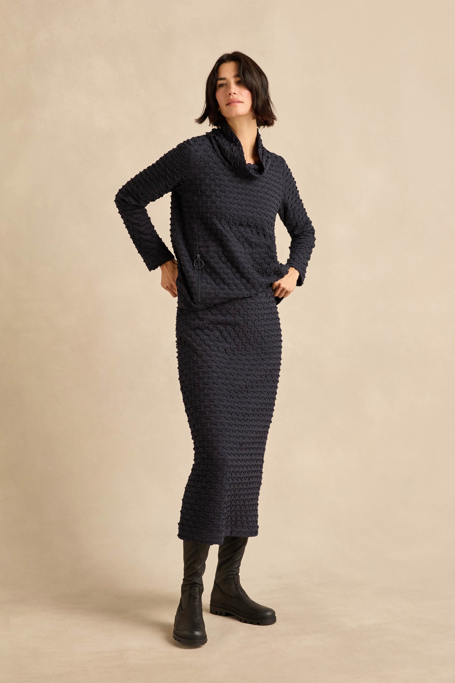 A woman stands against a plain beige background wearing the navy Pocket Jersey Tube Skirt and a matching textured stretch jersey long-sleeve top, paired with black boots.