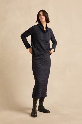 A woman stands against a plain beige background wearing the navy Pocket Jersey Tube Skirt and a matching textured stretch jersey long-sleeve top, paired with black boots.