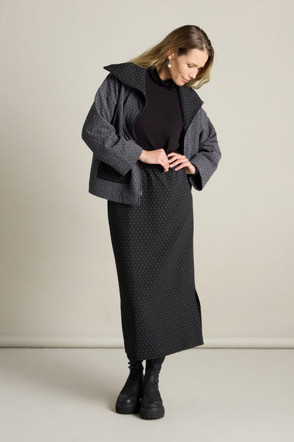 A person stands against a plain background, wearing a dark, long-sleeved jacket over the Square Jacquard Tube Skirt in an elegant tube silhouette, black boots, and a turtleneck sweater. The person gazes downwards.