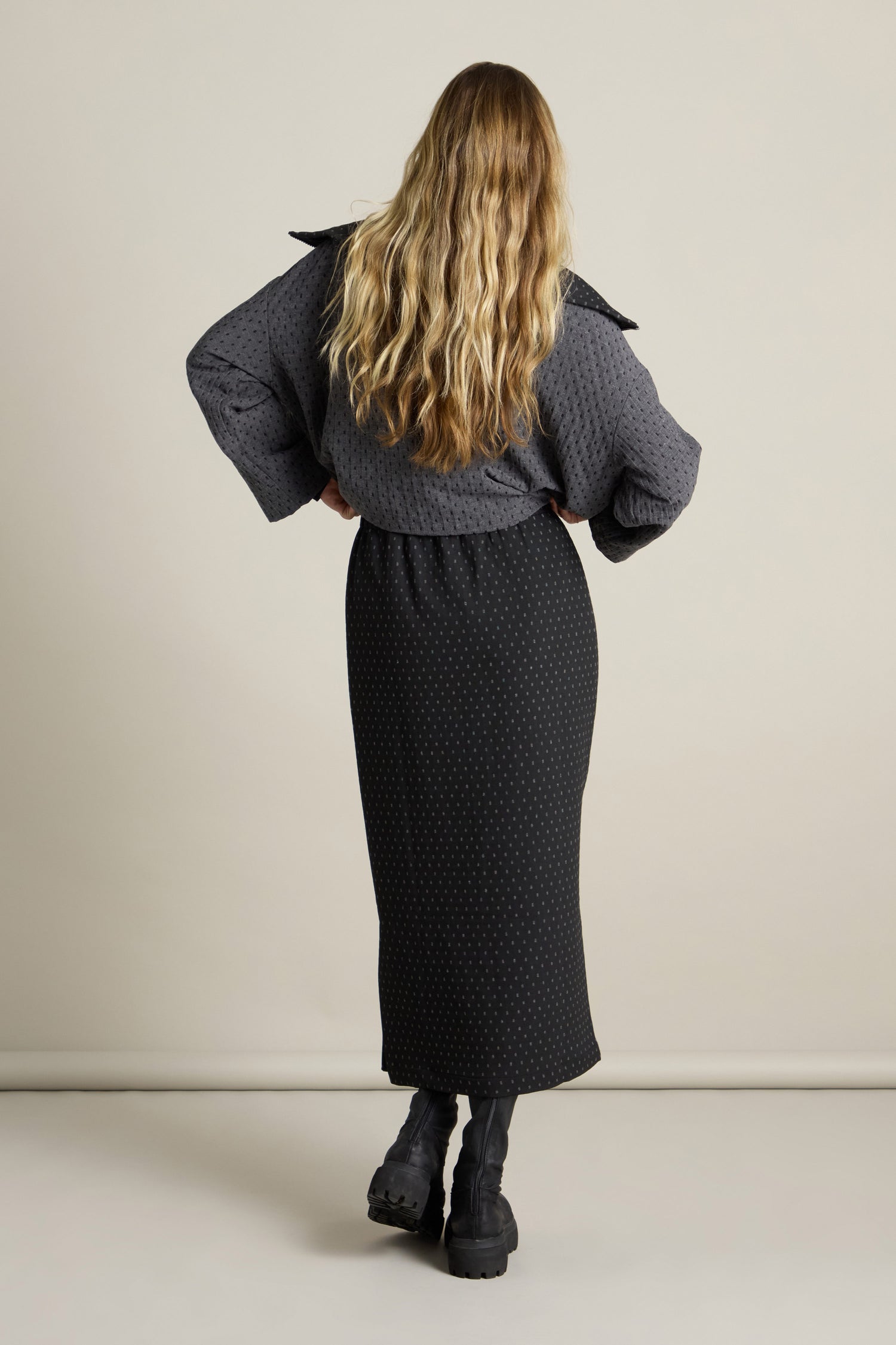 A woman with long blonde hair stands with her back to the camera, wearing a dark textured jacket and a Square Jacquard Tube Skirt in double-faced jacquard fabric, paired stylishly with black boots.