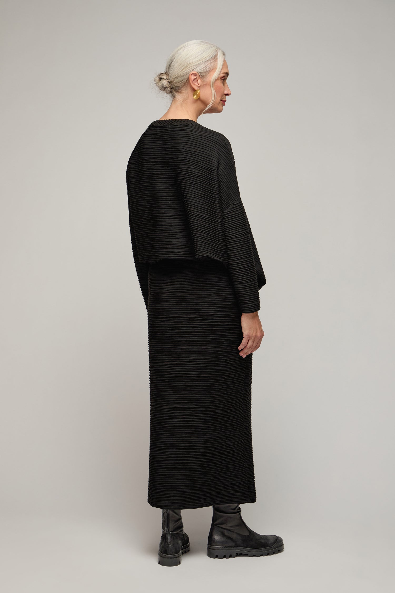 Woman with gray hair in a low bun wearing the Textured Wave Jersey Tube Skirt and black boots, standing sideways against a plain light background.