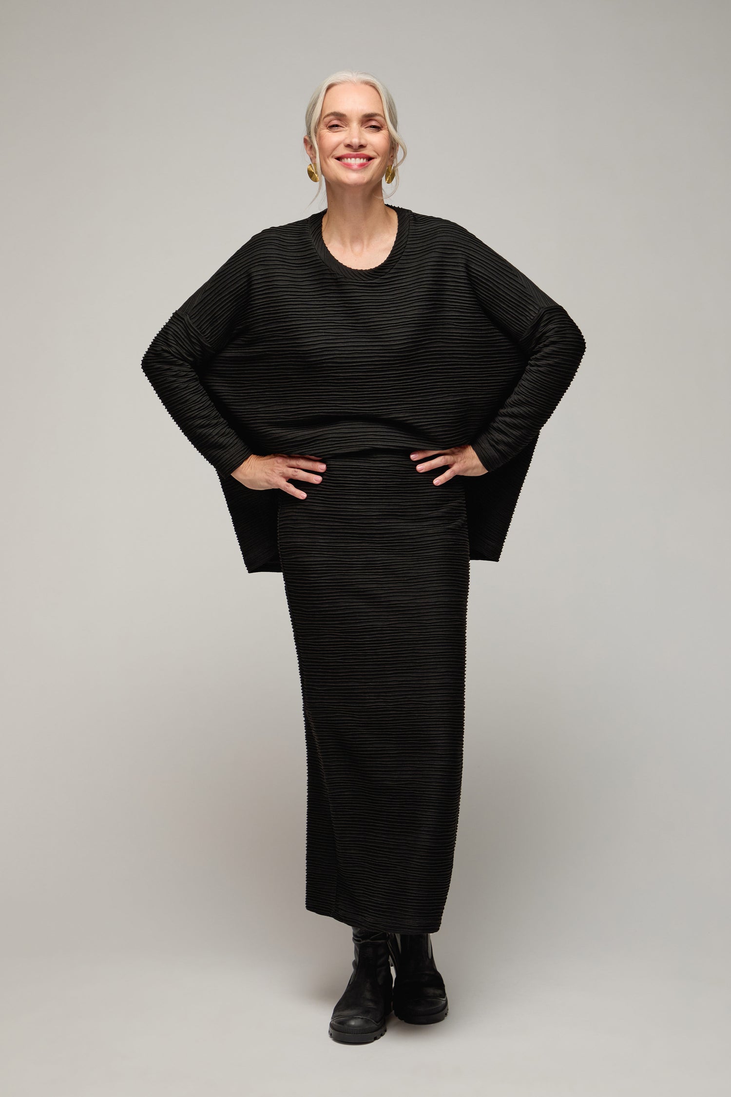 A woman with gray hair, wearing a black Textured Wave Jersey Tube Skirt and matching top with black boots, stands smiling with her hands on her hips against a plain background.