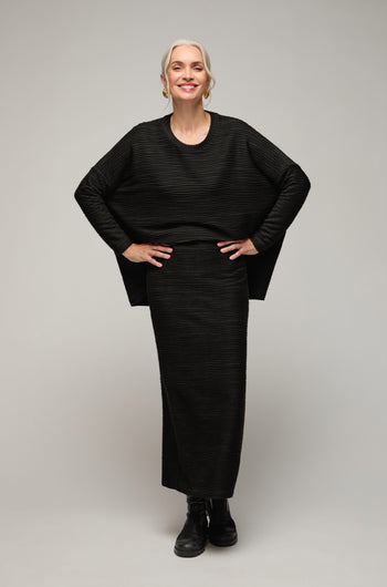 A woman with gray hair, wearing a black Textured Wave Jersey Tube Skirt and matching top with black boots, stands smiling with her hands on her hips against a plain background.