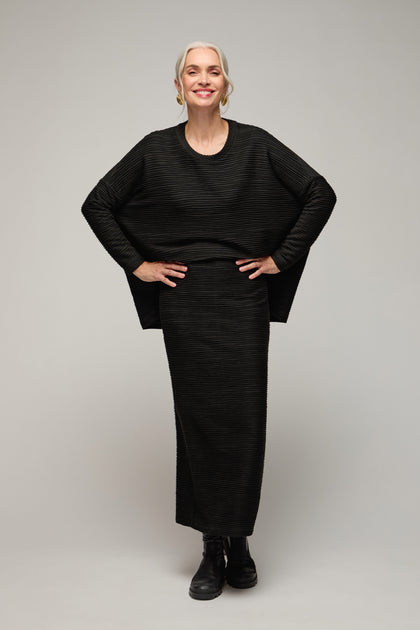 A woman with gray hair, wearing a black Textured Wave Jersey Tube Skirt and matching top with black boots, stands smiling with her hands on her hips against a plain background.
