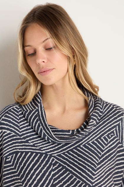 A woman with long blonde hair wears the Linear Prism Lines Cowl Neck Top in navy and white geometric print, made from soft brushed viscose, standing against a plain light background and looking down with a neutral expression.
