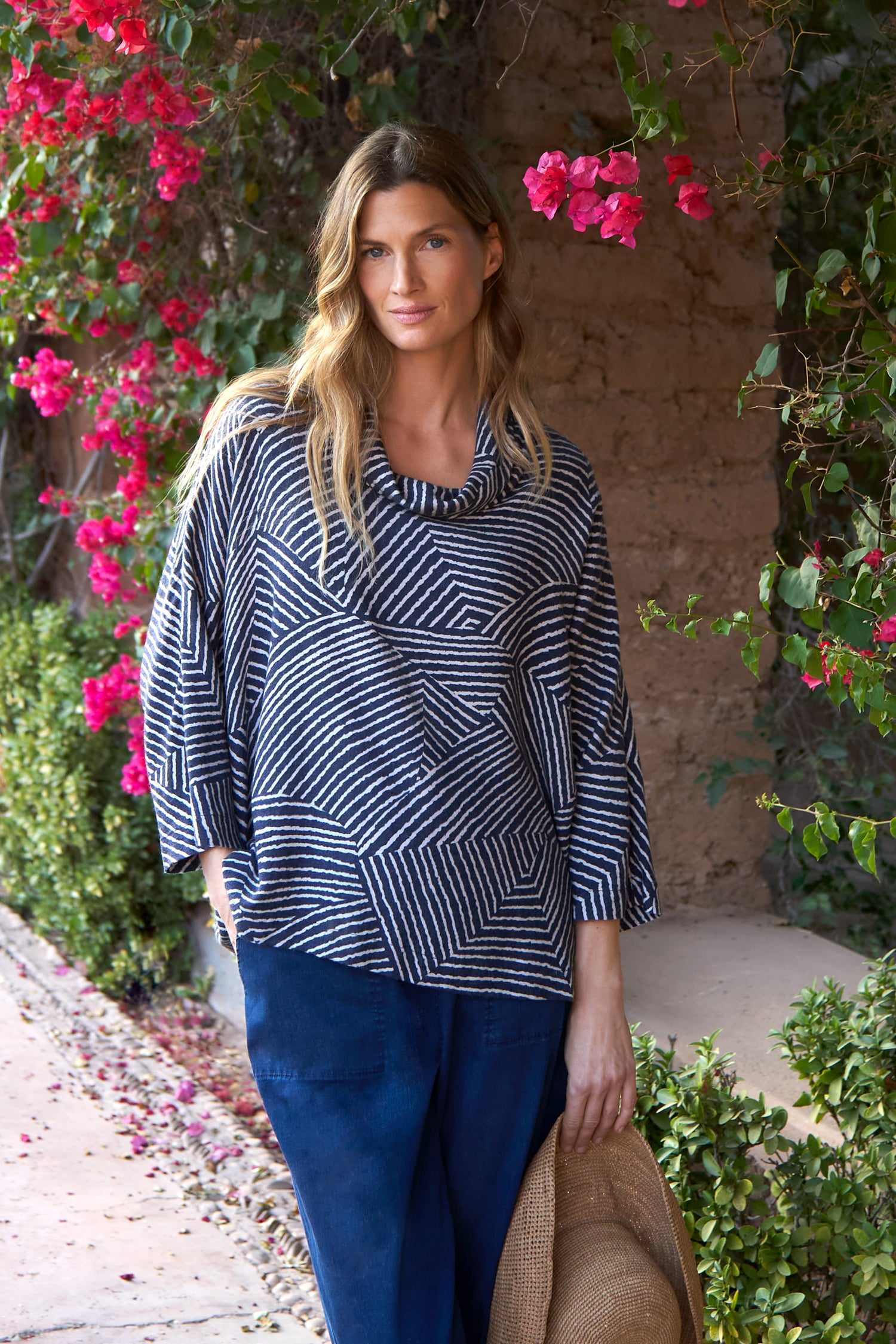 A woman stands outdoors by pink flowers and greenery, wearing the Linear Prism Cowl Neck Top and blue pants, holding a straw hat in one hand.