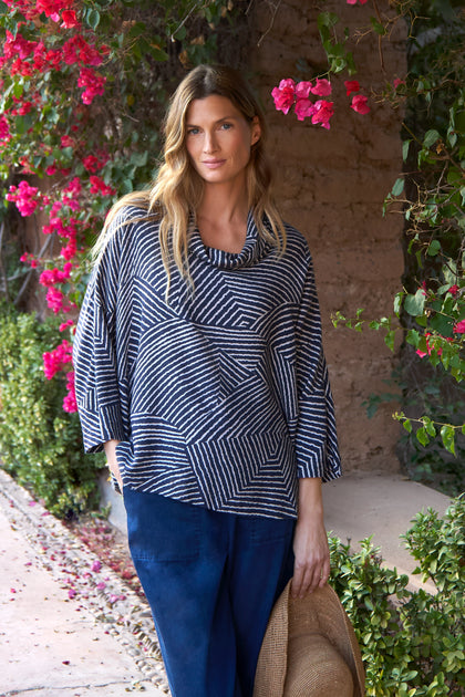 A woman stands outdoors by pink flowers and greenery, wearing the Linear Prism Cowl Neck Top and blue pants, holding a straw hat in one hand.