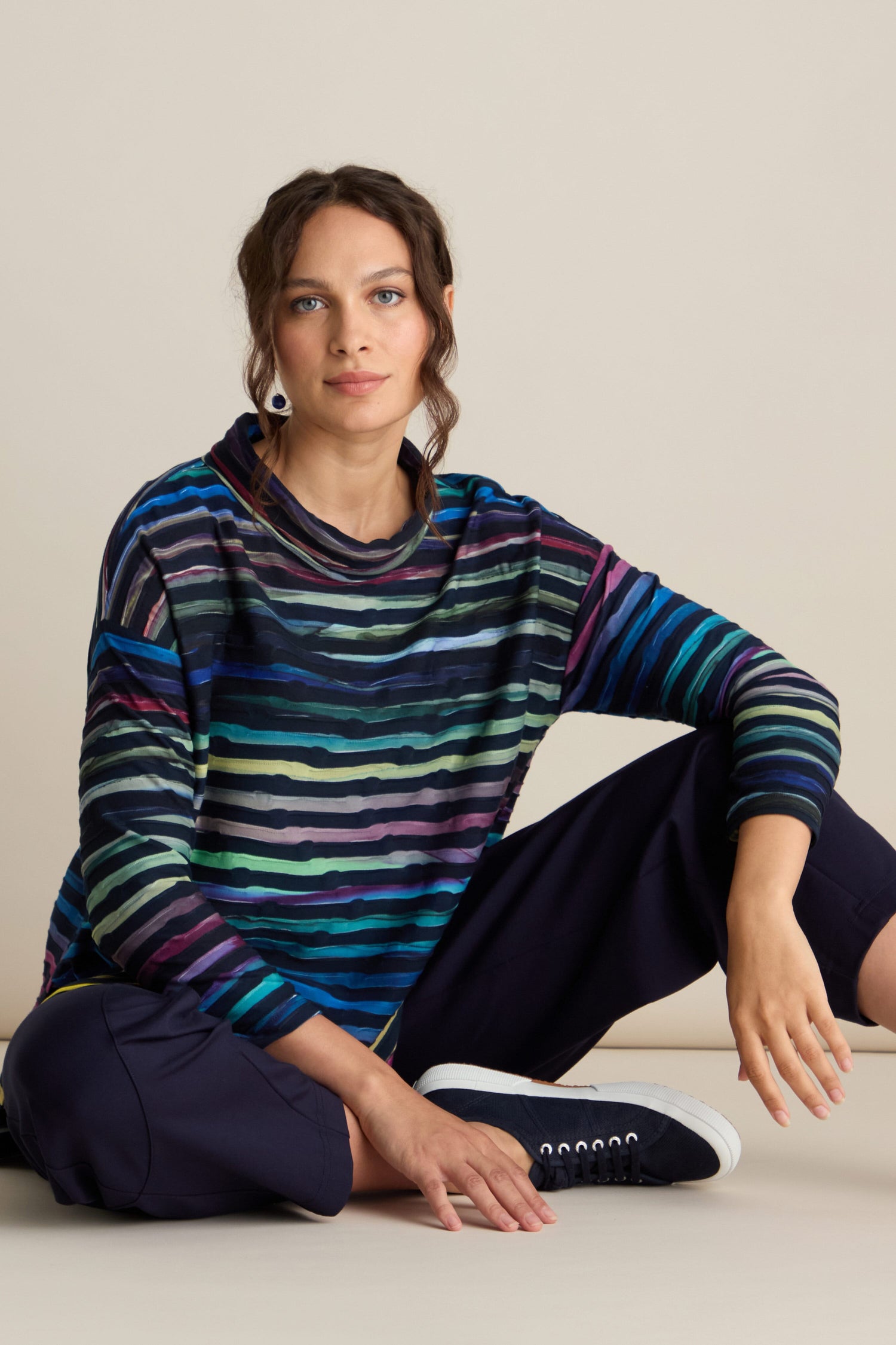 A person with long dark hair is sitting on the floor, dressed in wardrobe basics: the Textured Watercolour Landscape Top featuring colorful stripes, dark pants, and black and white sneakers. The unique textured jersey of their top adds flair as they look at the camera with a neutral expression.