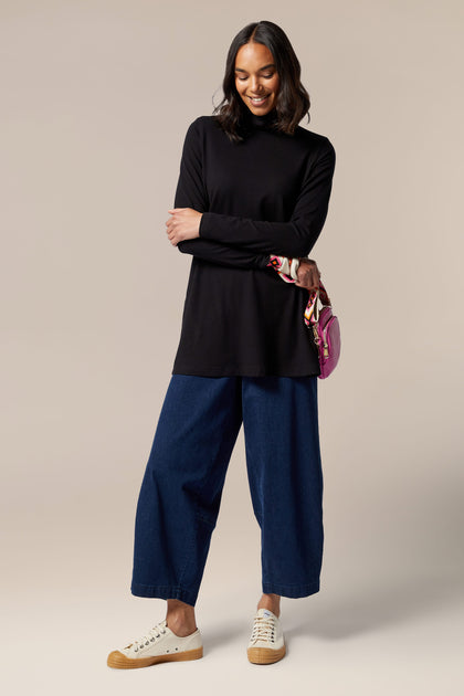 A woman wearing a black Fluid Crepe Flared Polo Neck top and blue jeans.