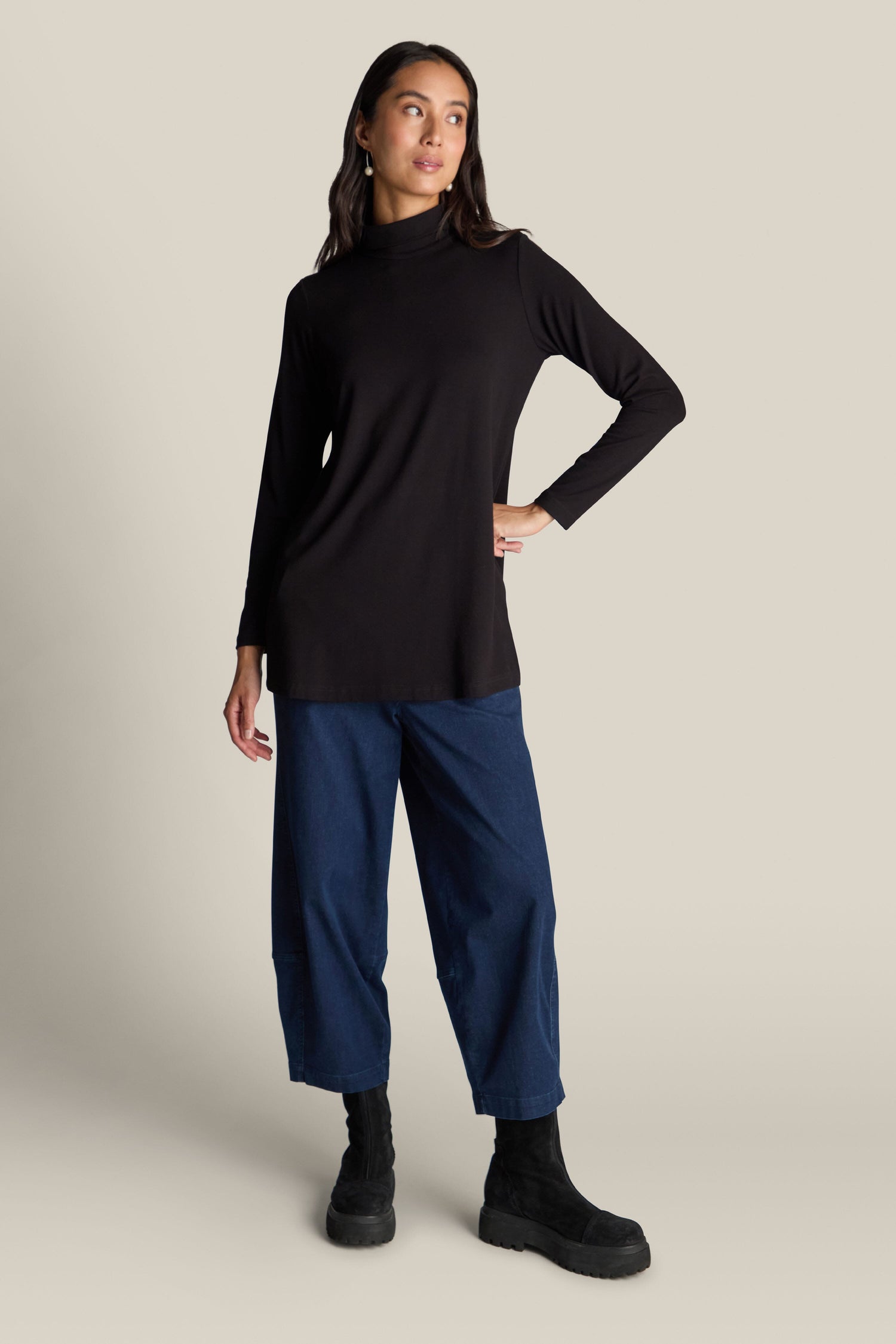 A person stands in a relaxed pose, wearing the Fluid Crepe Flared Polo Neck in a long-sleeved black turtleneck style, blue wide-leg pants, and black boots, against a plain beige background.