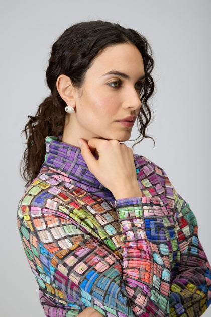 A woman with dark curly hair poses in profile, wearing the vibrant Coloured Glass Tiles Jersey Tunic with a hand near her chin against a plain background.