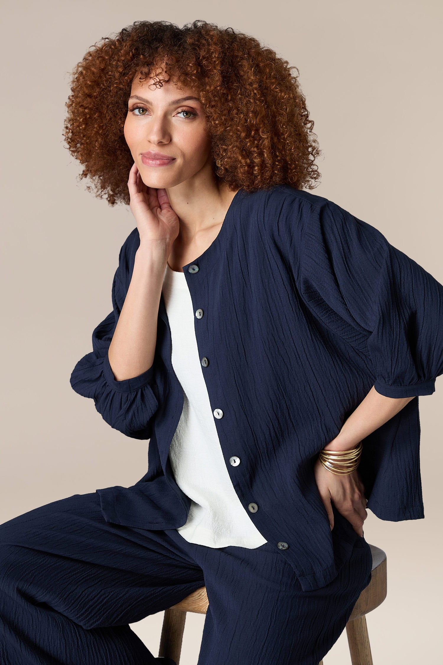 The woman is wearing a navy pleated Crinkle Soft Viscose Shirt and pants while sitting on a stool.
