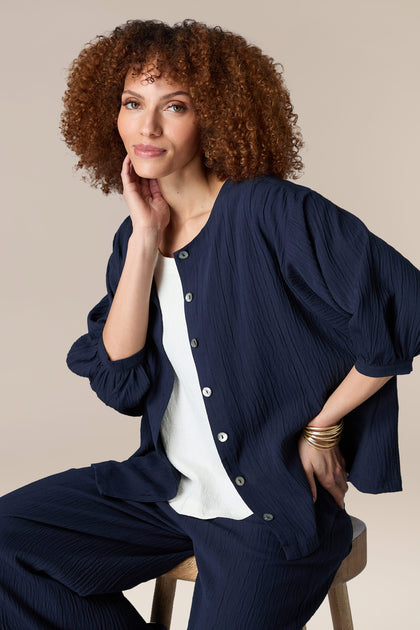 The woman is wearing a navy pleated Crinkle Soft Viscose Shirt and pants while sitting on a stool.