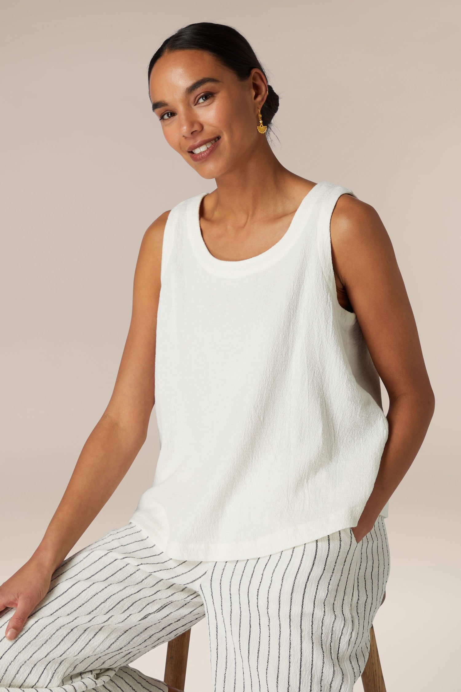 A woman in a white sleeveless top and a Linen Viscose Vest, along with striped pants, is sitting on a wooden stool against a neutral backdrop, smiling softly towards the camera.