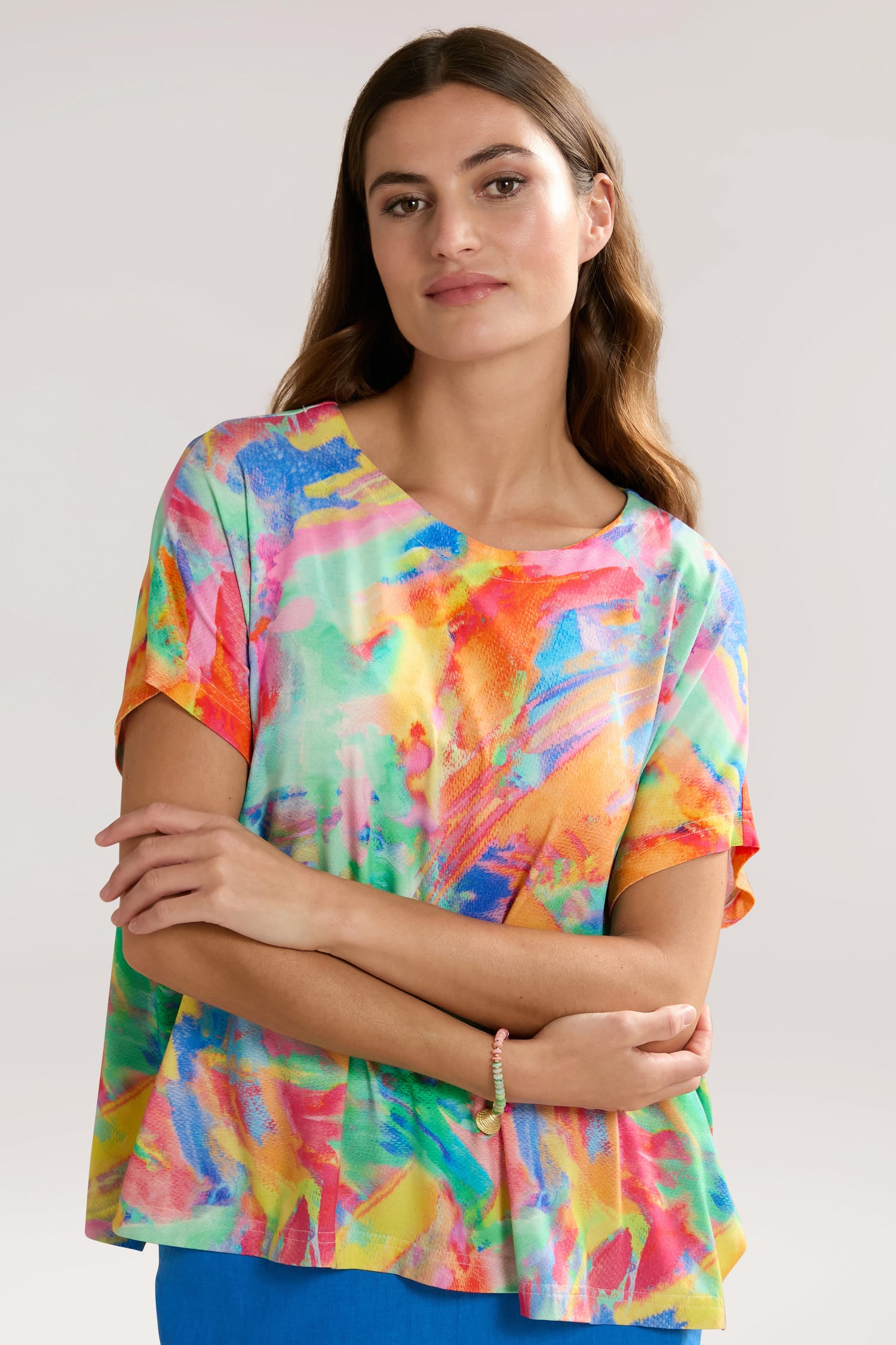 A woman wearing an elegant, colorful abstract-patterned Vibrant Watercolour Jersey Top stands with her arms crossed against a plain background.