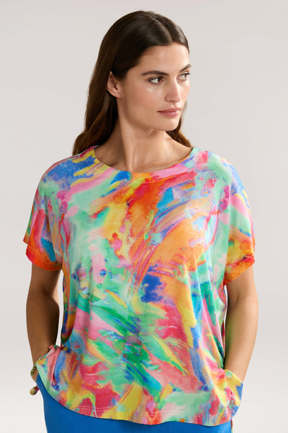 A woman in a colorful Vibrant Watercolour Jersey Top with abstract patterns stands with her hands in her pockets, looking to the side. She has long brown hair and a neutral expression. The background is plain light gray, complementing her elegant style crafted from sustainable LENZING™ Viscose ECOVERO™.