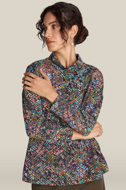 A woman with her hair up, wearing the stylish Diamond Jacquard Jersey Top made from multi-colored jacquard fabric, poses with her arms crossed against a plain background.