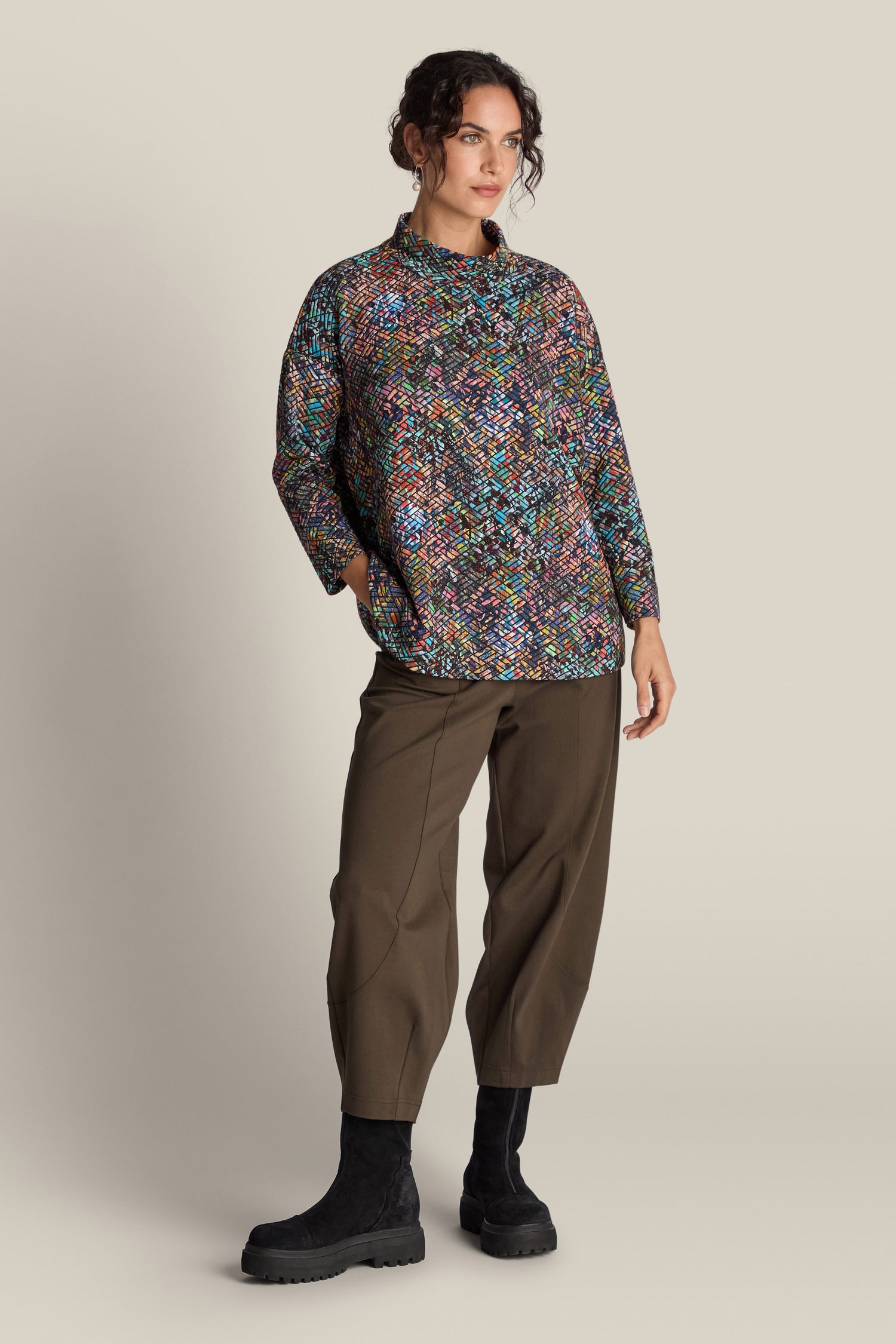 A person stands against a plain background, wearing the Diamond Jacquard Jersey Top in colorful patterns, paired with olive green pants and black boots.