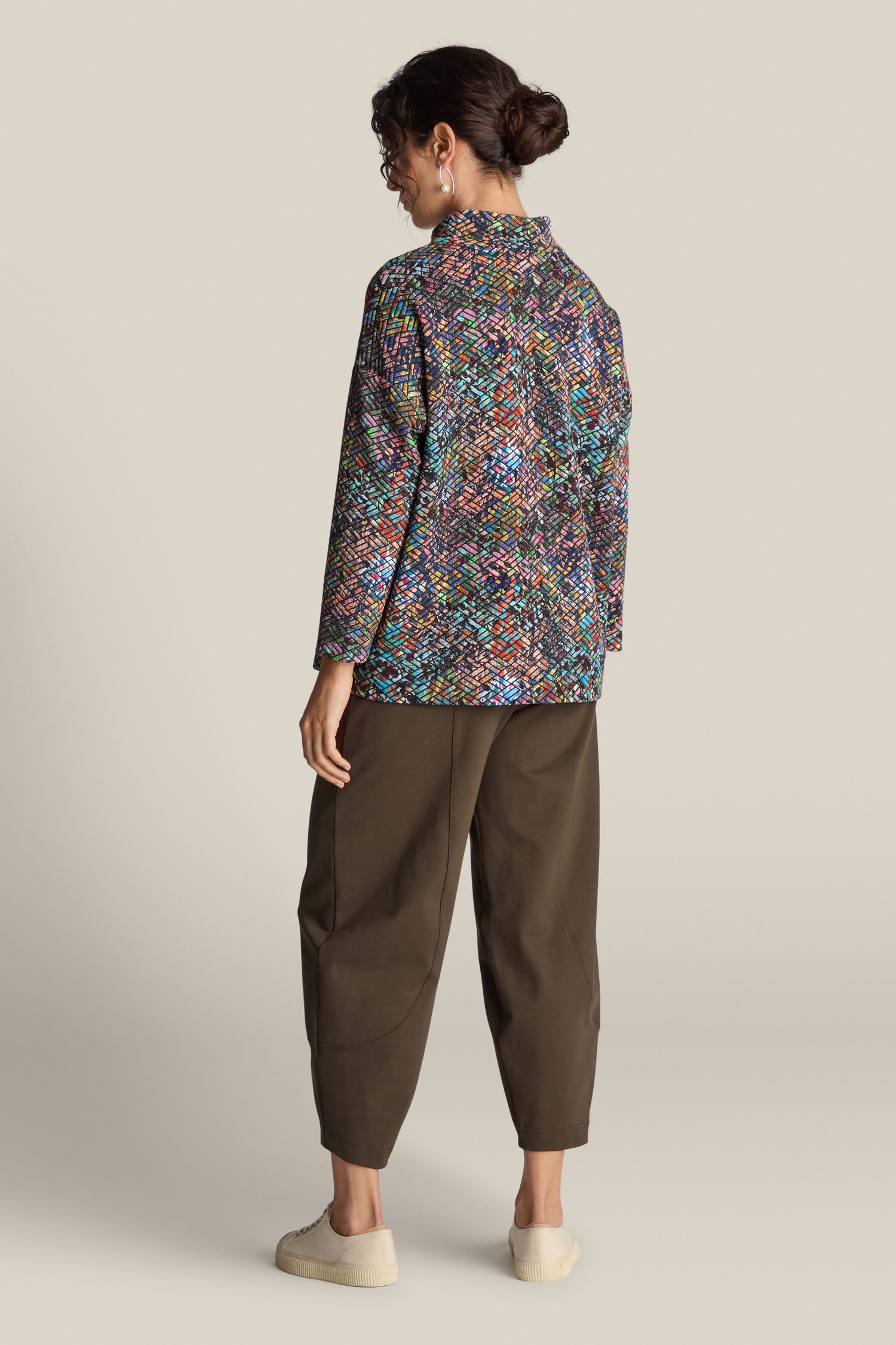 A person with dark hair styled in a bun wears the Diamond Jacquard Jersey Top, which features a vibrant multi-colored diamond pattern. They are also dressed in olive green pants and white sneakers, standing with their back turned against a plain beige background.