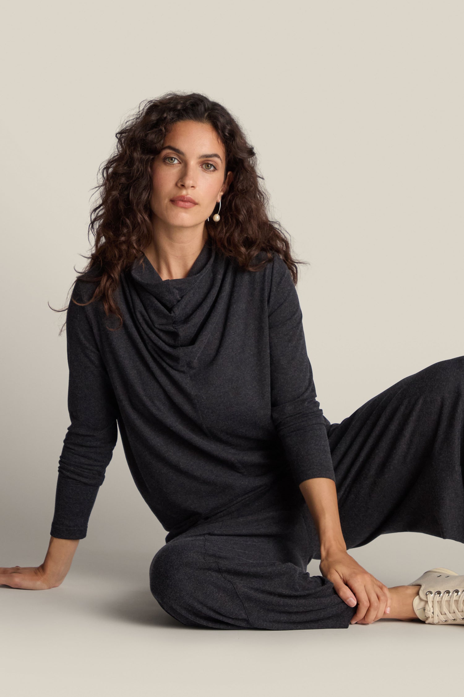 A person with long curly hair, dressed in a Soft Viscose Jersey Cowl Neck Top and matching pants, sits in a relaxed pose against a plain background, exuding elegance and comfort.