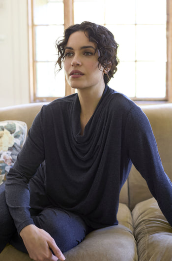 A person with short curly hair is sitting on a beige sofa, elegantly dressed in a dark, long-sleeve Soft Viscose Jersey Cowl Neck Top, with a neutral expression.