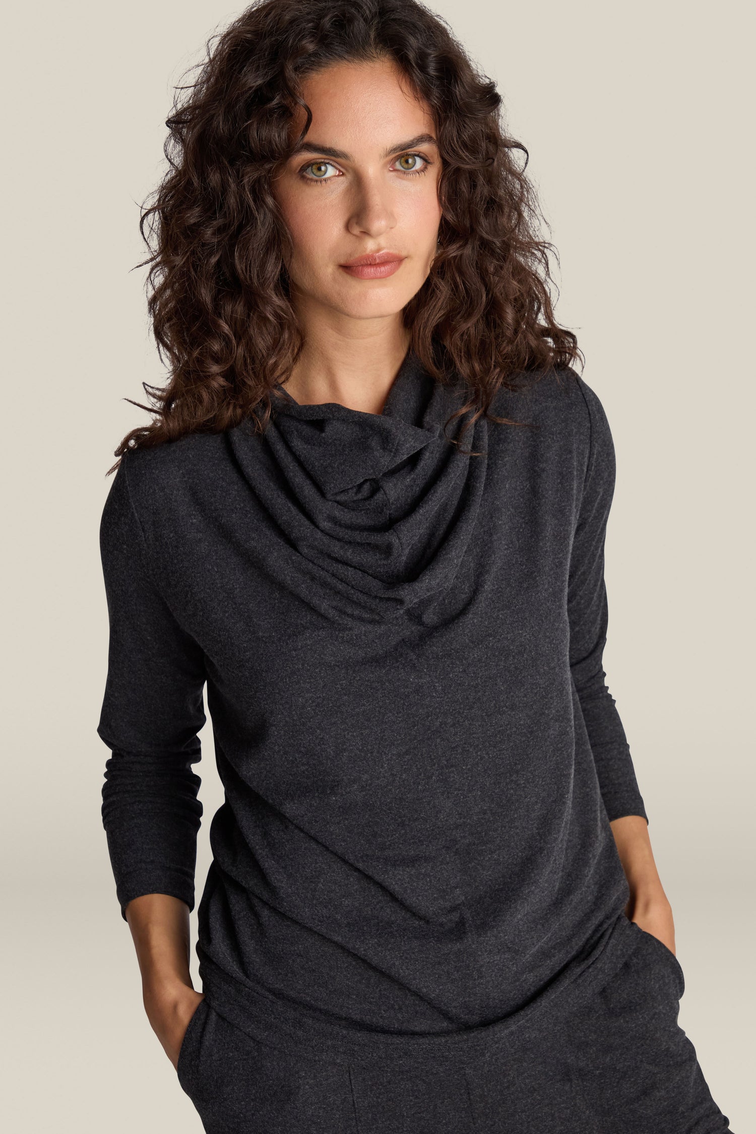 A woman with curly hair wearing the Soft Viscose Jersey Cowl Neck Top in dark gray stands against a plain background, exuding both elegance and comfort.