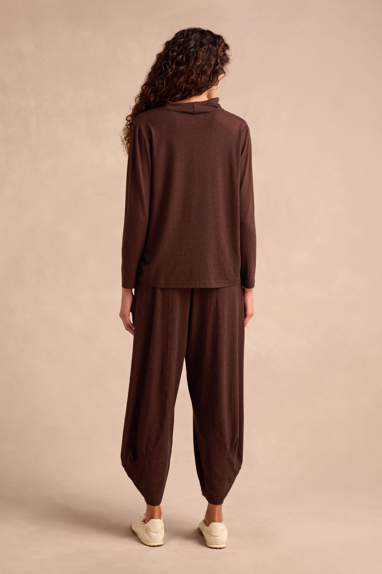 Person standing against a plain background, wearing the Soft Viscose Jersey Ruched Top in brown—a loose-fitting, long-sleeve cowl neck style—paired with matching wide-leg pants and white sneakers, facing away from the camera.
