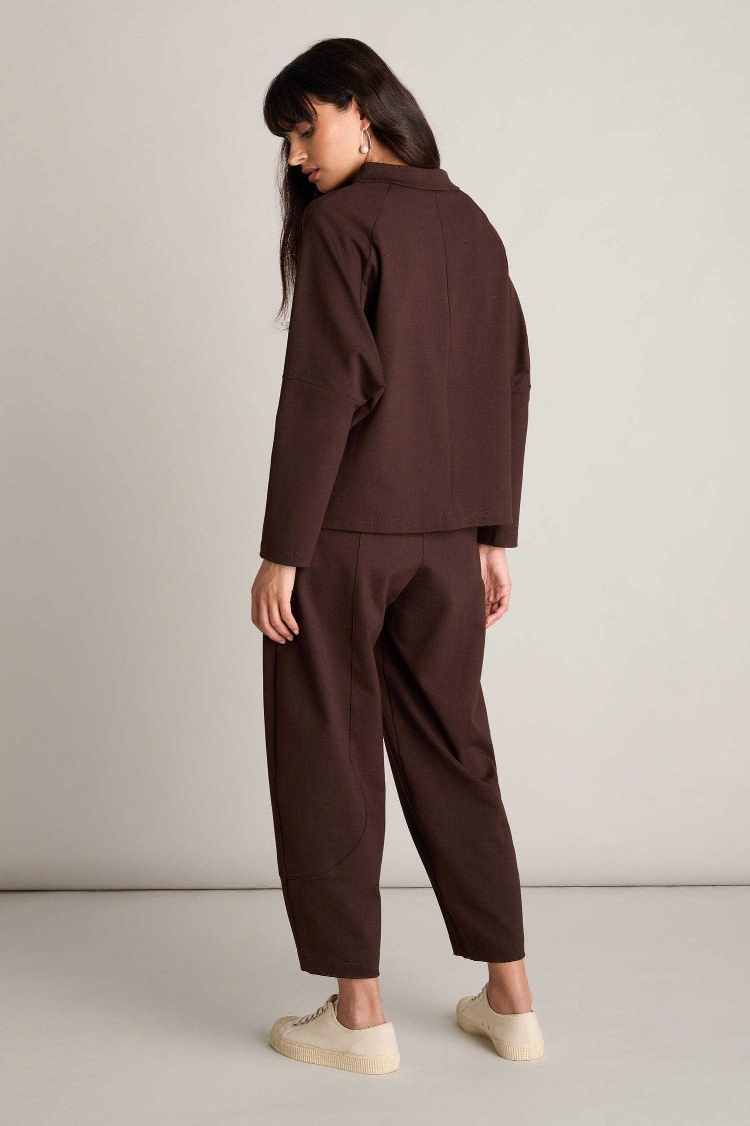 A woman with long dark hair, styled in a casual chic manner, stands sideways wearing the brown Ponte Boxy Top and matching pants, accompanied by white sneakers. Her gaze is directed downward.