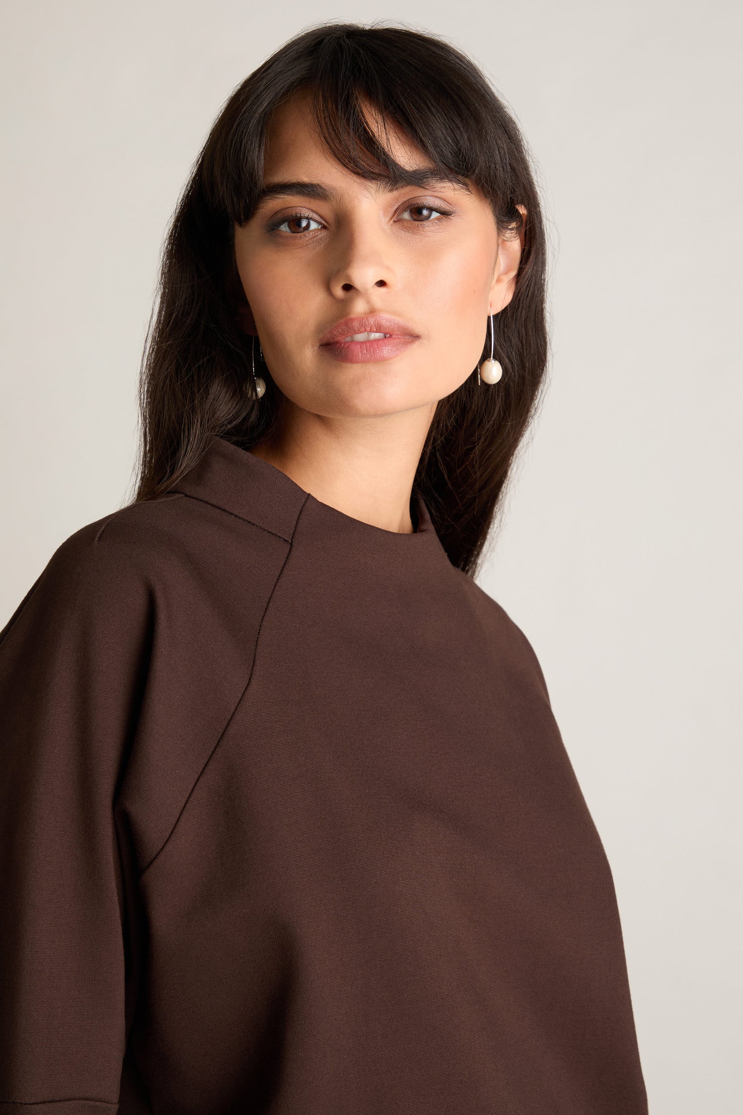 A person with long dark hair wearing the Ponte Boxy Top and pearl drop earrings looks at the camera against a plain background, effortlessly showcasing a casual chic look.