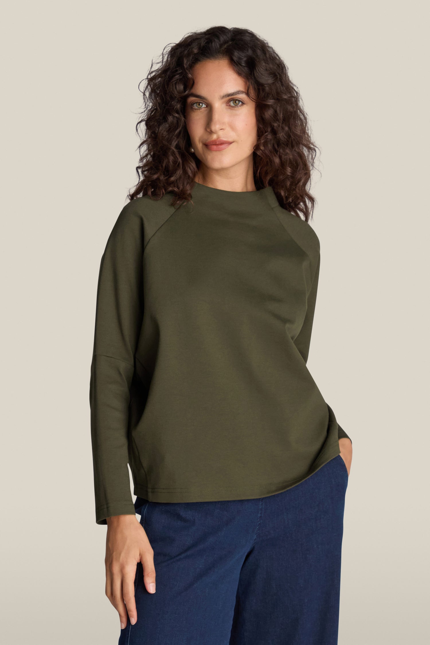 A person with curly hair wearing a long-sleeve olive green Ponte Boxy Top and blue jeans effortlessly stands against a plain background.