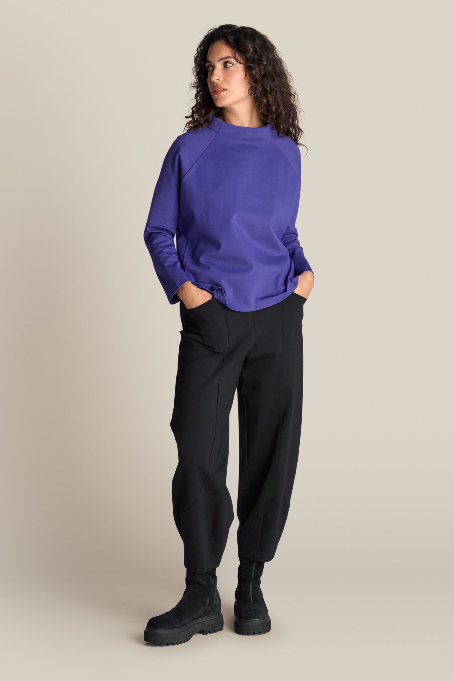 A woman stands against a plain background, exuding a contemporary edge with her Ponte Boxy Top in purple, black pants, and black boots, her hands casually in her pockets.