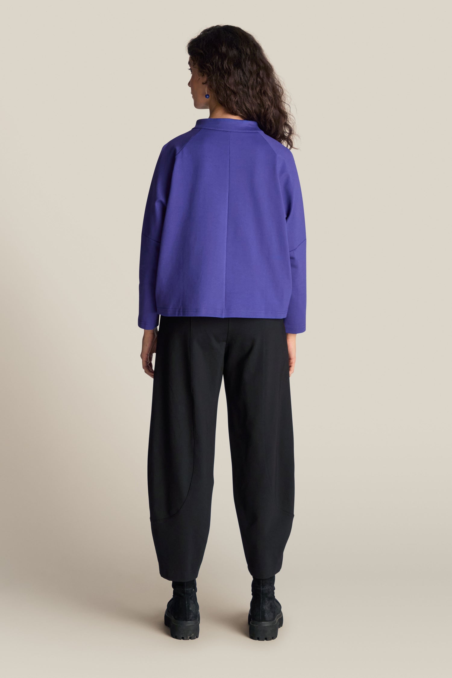 A person with curly hair wearing the Ponte Boxy Top in purple and black pants, viewed from the back, standing against a plain background, exuding a casual chic look with a contemporary edge.