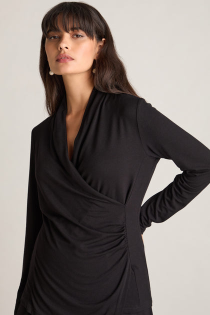 A woman with long hair, wearing a Fluid Crepe Jersey Ruched Side Top and pearl earrings, stands with one hand on her hip, highlighting her hourglass silhouette as she faces the camera.