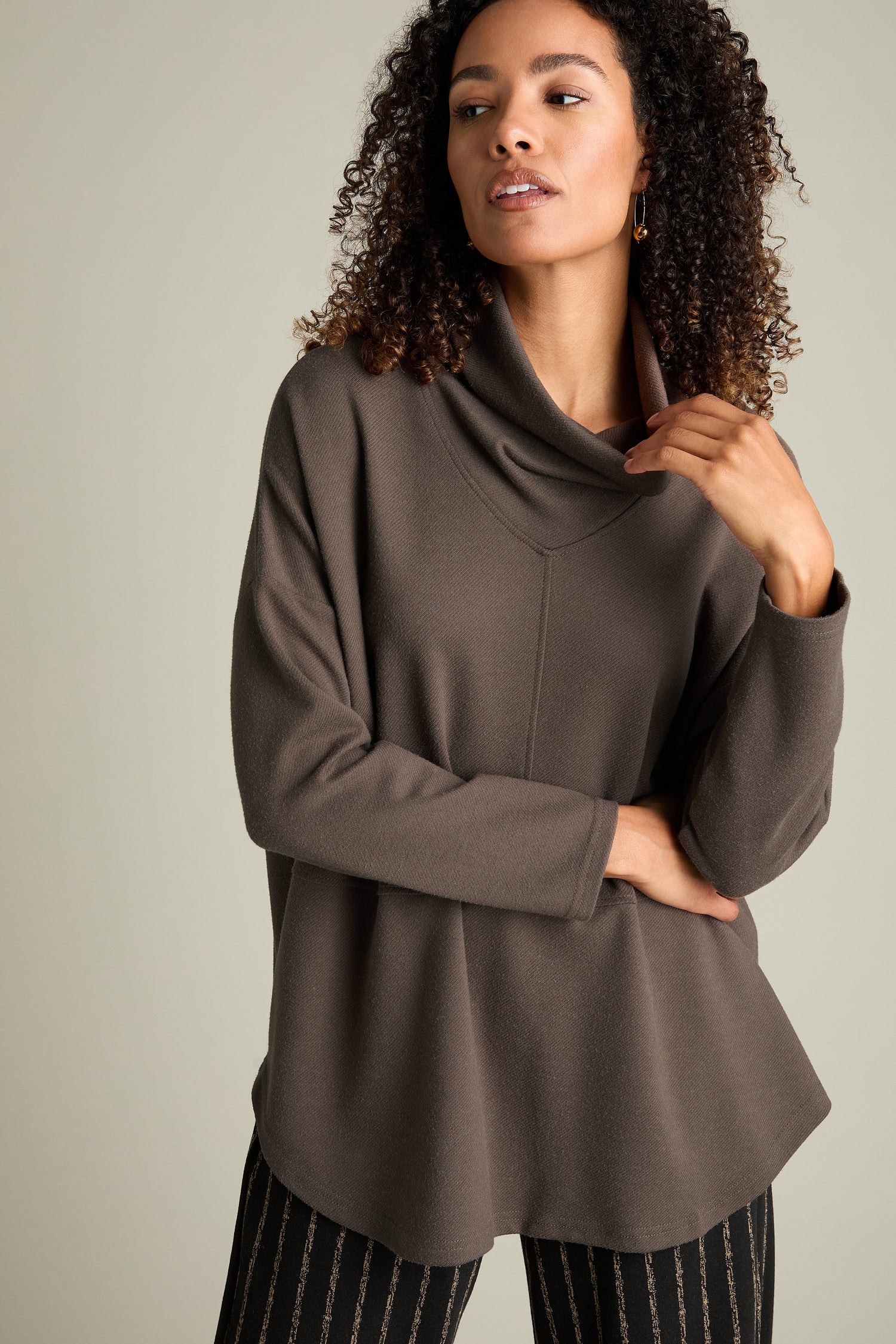 A woman with curly hair stands against a gray background, wearing the Marl Jersey Sweatshirt, a loose, long-sleeve brown top featuring a diagonal marl knit pattern, paired with striped pants.