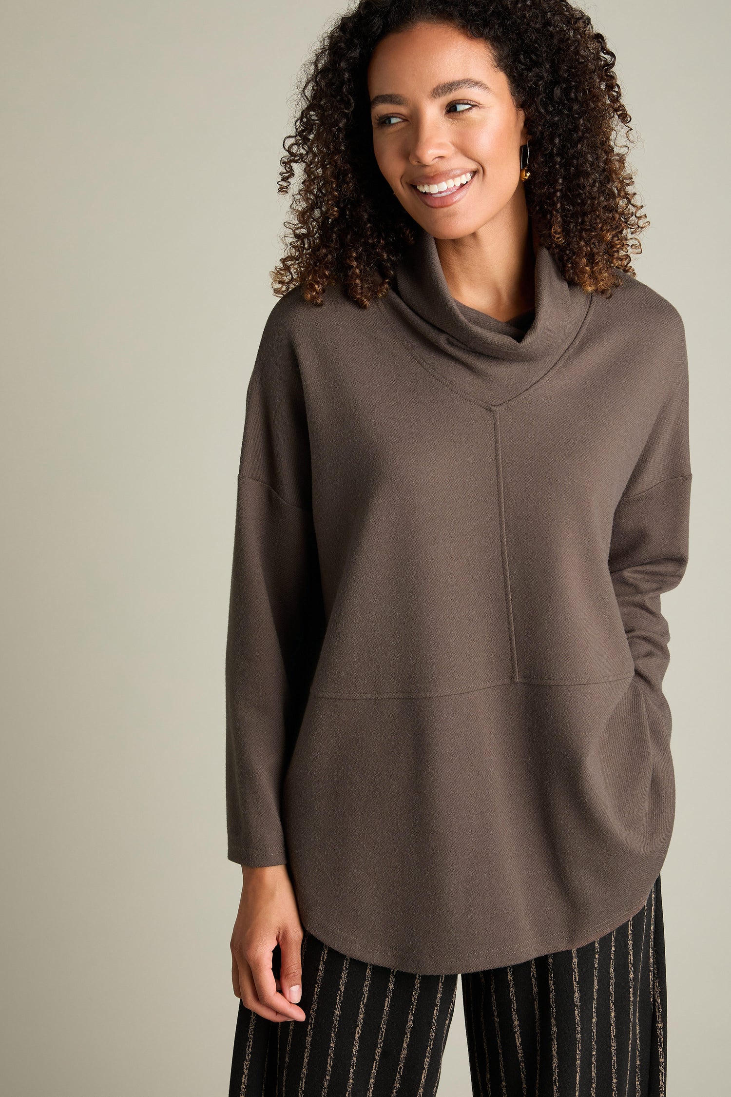 A person with curly hair smiles while wearing the Marl Jersey Sweatshirt, a brown long-sleeve top with a funnel cowl neck, paired with striped pants, standing against a plain background.