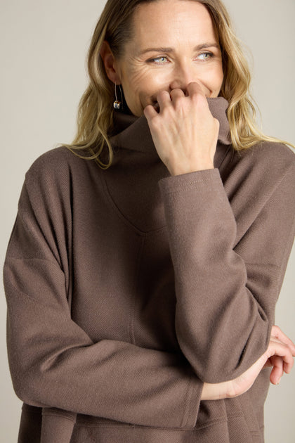 A person with long hair is smiling and covering part of their face with their hand while wearing the Marl Jersey Sweatshirt in brown.