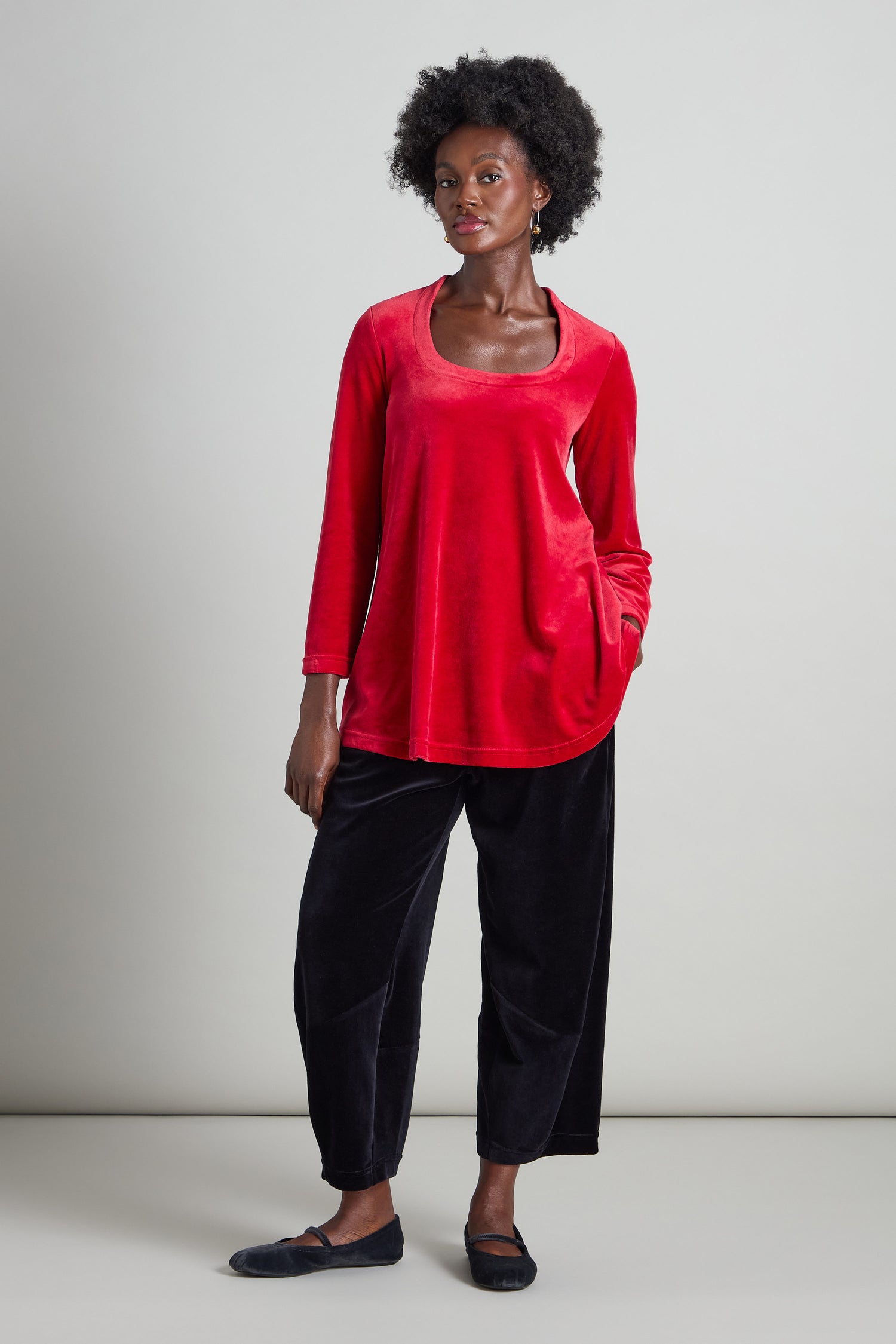 A person wearing a Velvet Jersey Horseshoe Neck Top in red, featuring a fit-and-flare silhouette, paired with black pants, stands against a plain background.