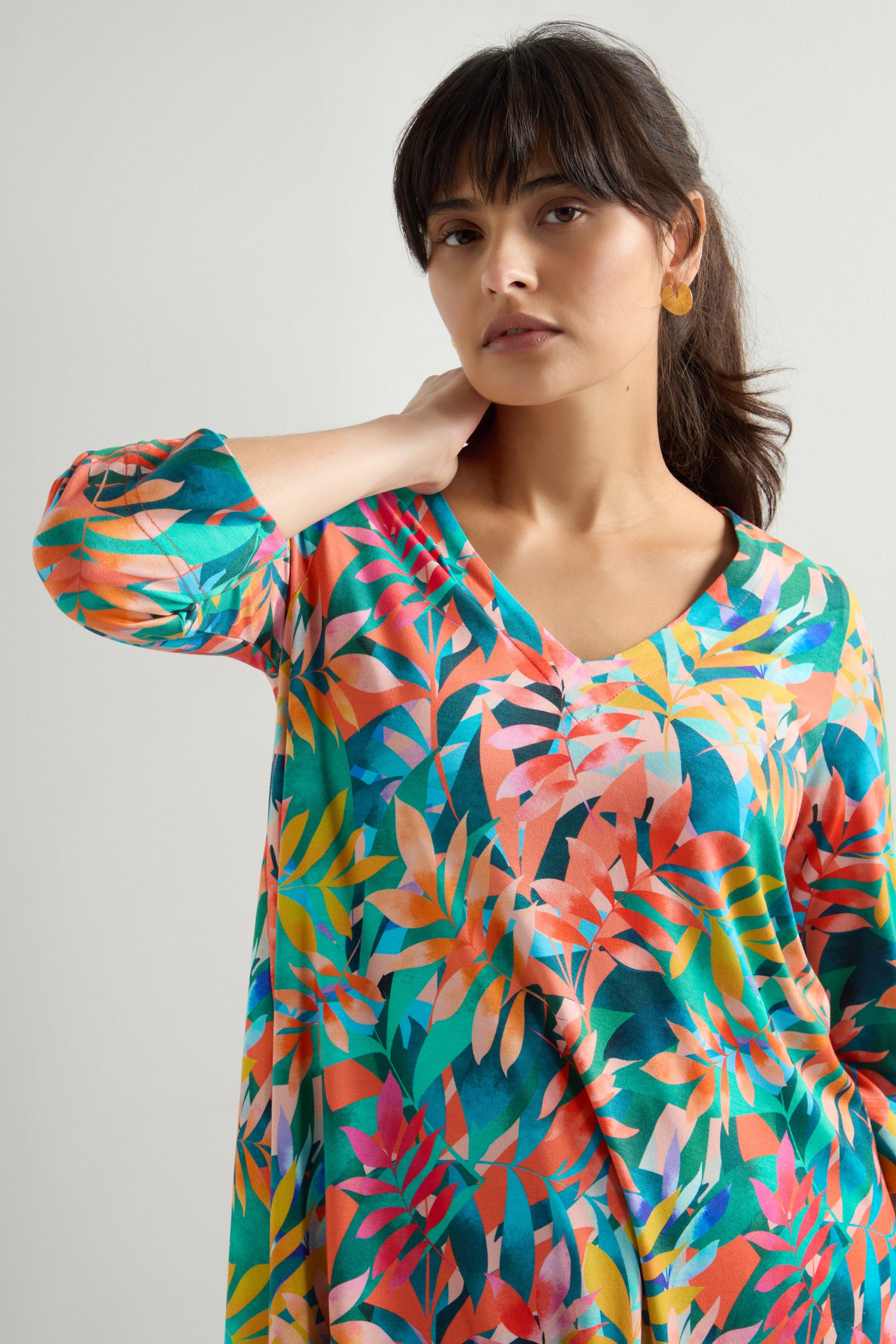 A person in a Tropical Leaves Jersey Print Tunic stands against a neutral background, one hand touching their neck.